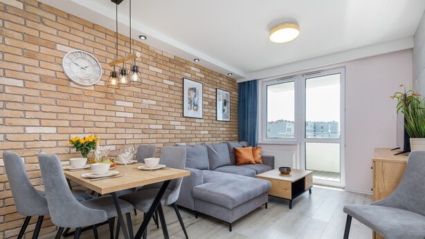 A modern living room with a brick wall, a grey sofa, a wooden dining table with chairs, and a large window with a view. - Seaside Apartamenty Platany Kołobrzeg  (Kolobrzeg)