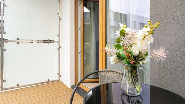 Una mesa de vidrio con un florero de flores en un balcón con piso de madera y puerta corrediza de vidrio.