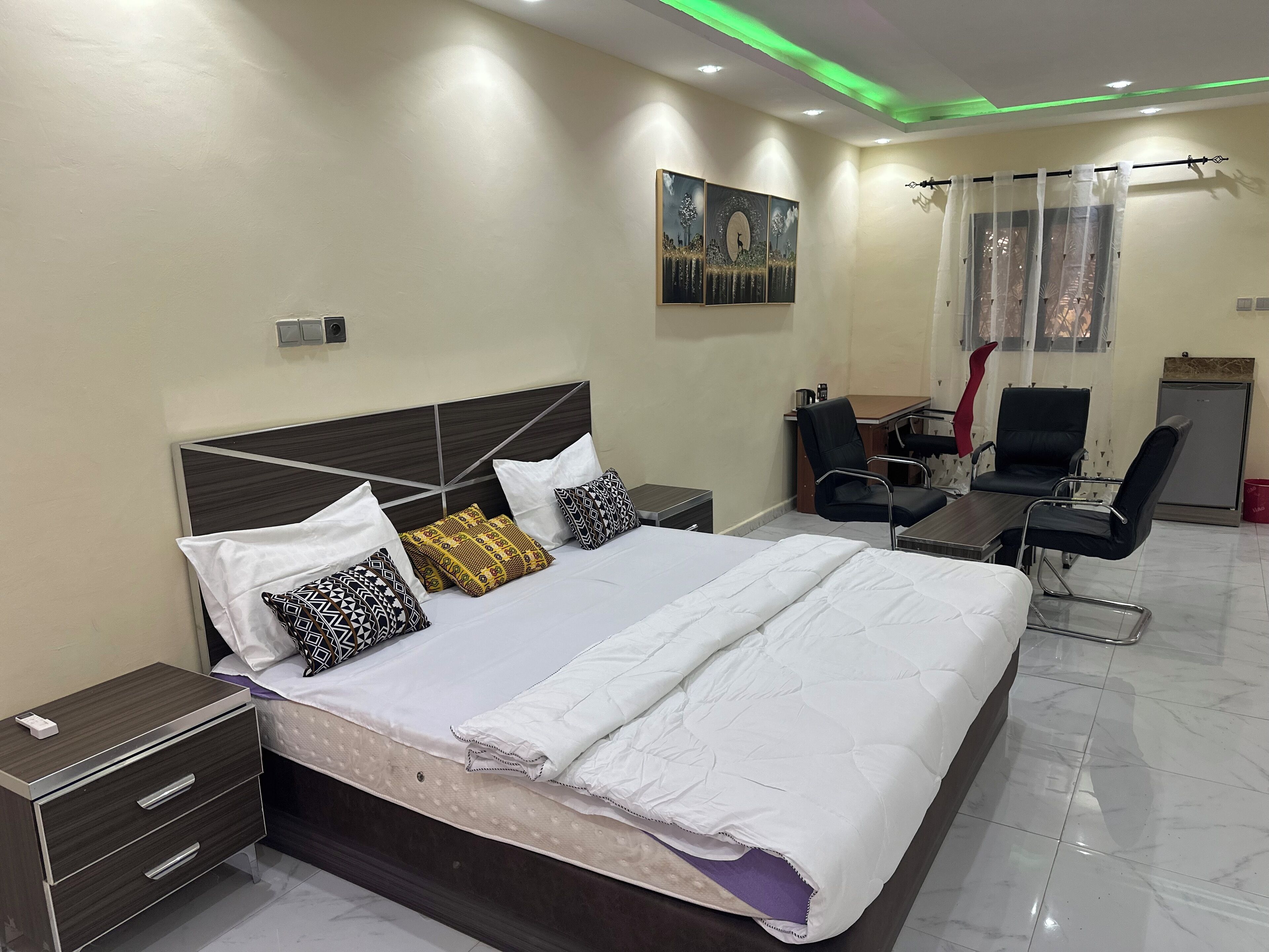 A hotel room with a large bed, bedside tables, a desk, two chairs, a small refrigerator, and a painting on the wall.