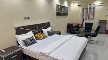 A hotel room with a large bed, bedside tables, a desk, two chairs, a small refrigerator, and a painting on the wall.