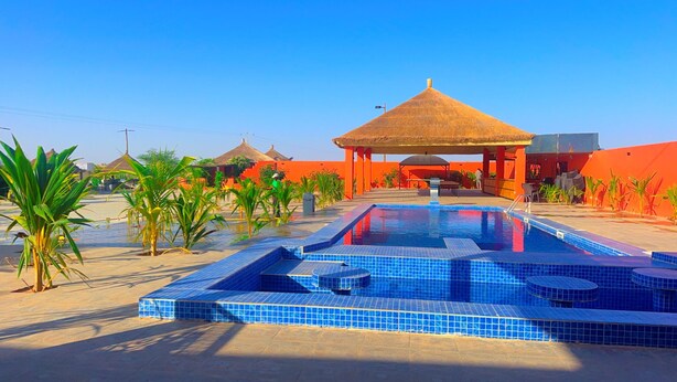 Una piscina con techo de paja, rodeada de palmeras y un edificio rojo.
