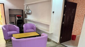 Una habitación con dos sillones morados, una mesa de centro de madera, un estante blanco, una puerta oscura y un cubo rojo en el suelo.
