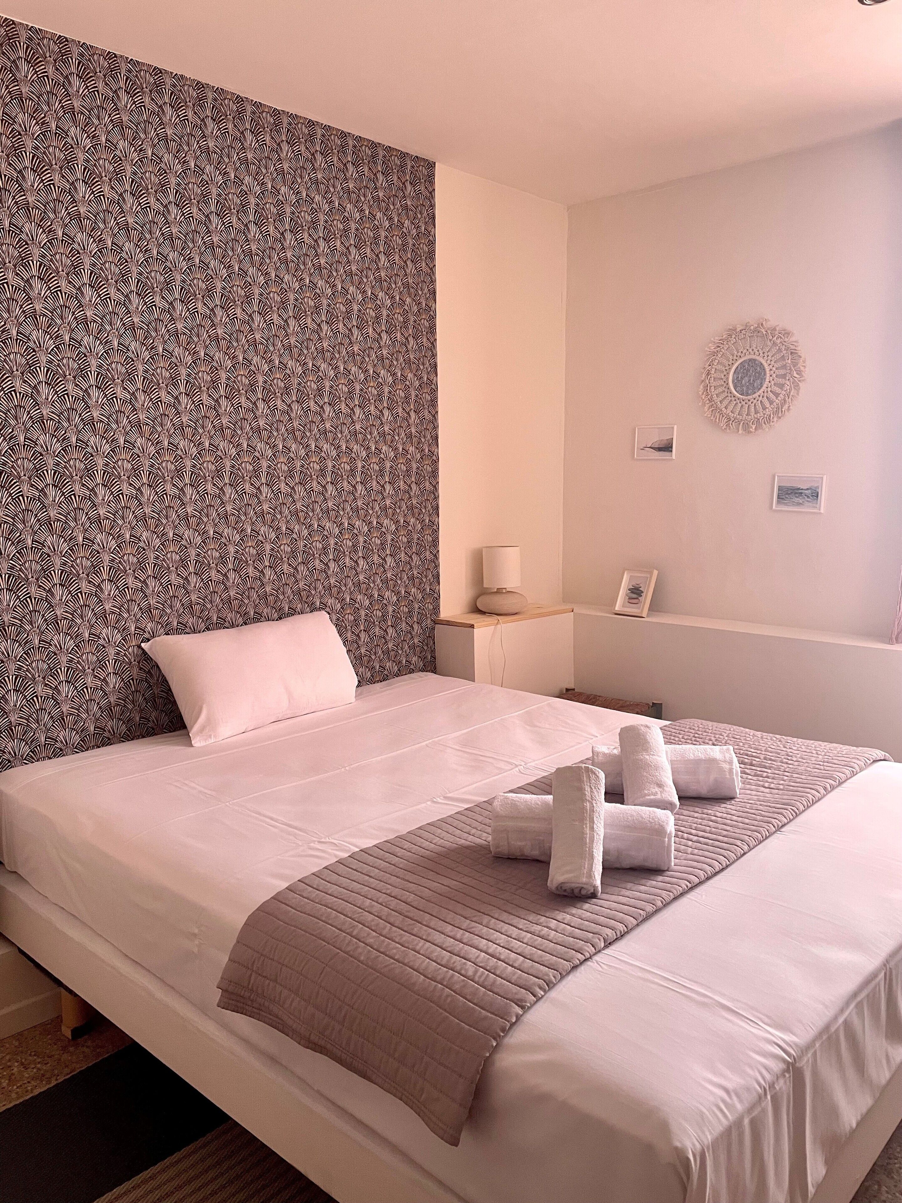 A neatly made bed with a patterned headboard, a bedside table with a lamp, and a wall with a macrame wall hanging.