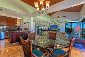A spacious living area with a glass dining table surrounded by wicker chairs, a kitchen with wooden cabinets, and a large window offering a view of the outdoors. - Exquisite Prime Beachfront Haven, Indulge in Tropical Luxury -Wailea Elua 1302 (Wailea, Maui)