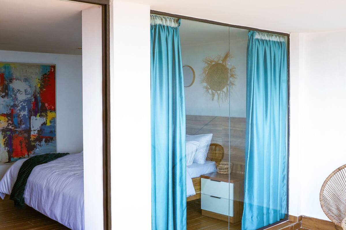 A bedroom with a bed, a white bedside table, a wicker basket, and a blue curtain.