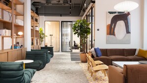 A modern office space with a long row of green sofas, a brown sofa, and a white table. There are bookshelves on the left and a large painting on the right wall. - Noli Otaniemi (Espoo)
