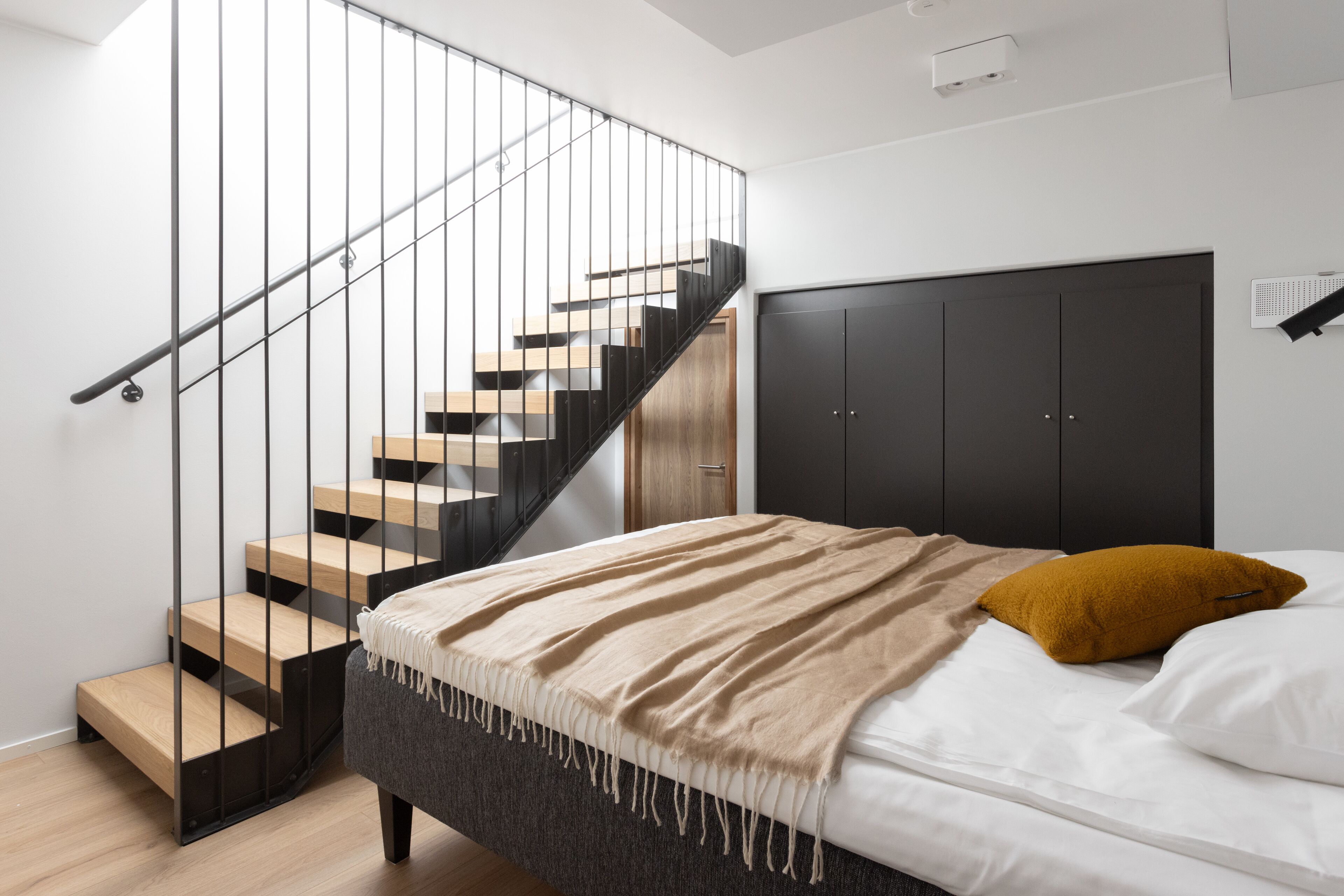 A modern bedroom with a staircase, a bed with white bedding and a mustard pillow, and a dark headboard.
