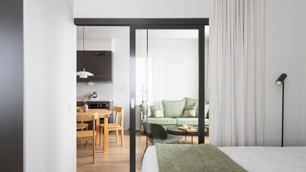 A modern hotel room with a bed, a dining area with a table and chairs, a sofa, and a sliding door leading to a balcony. - Noli Otaniemi (Espoo)