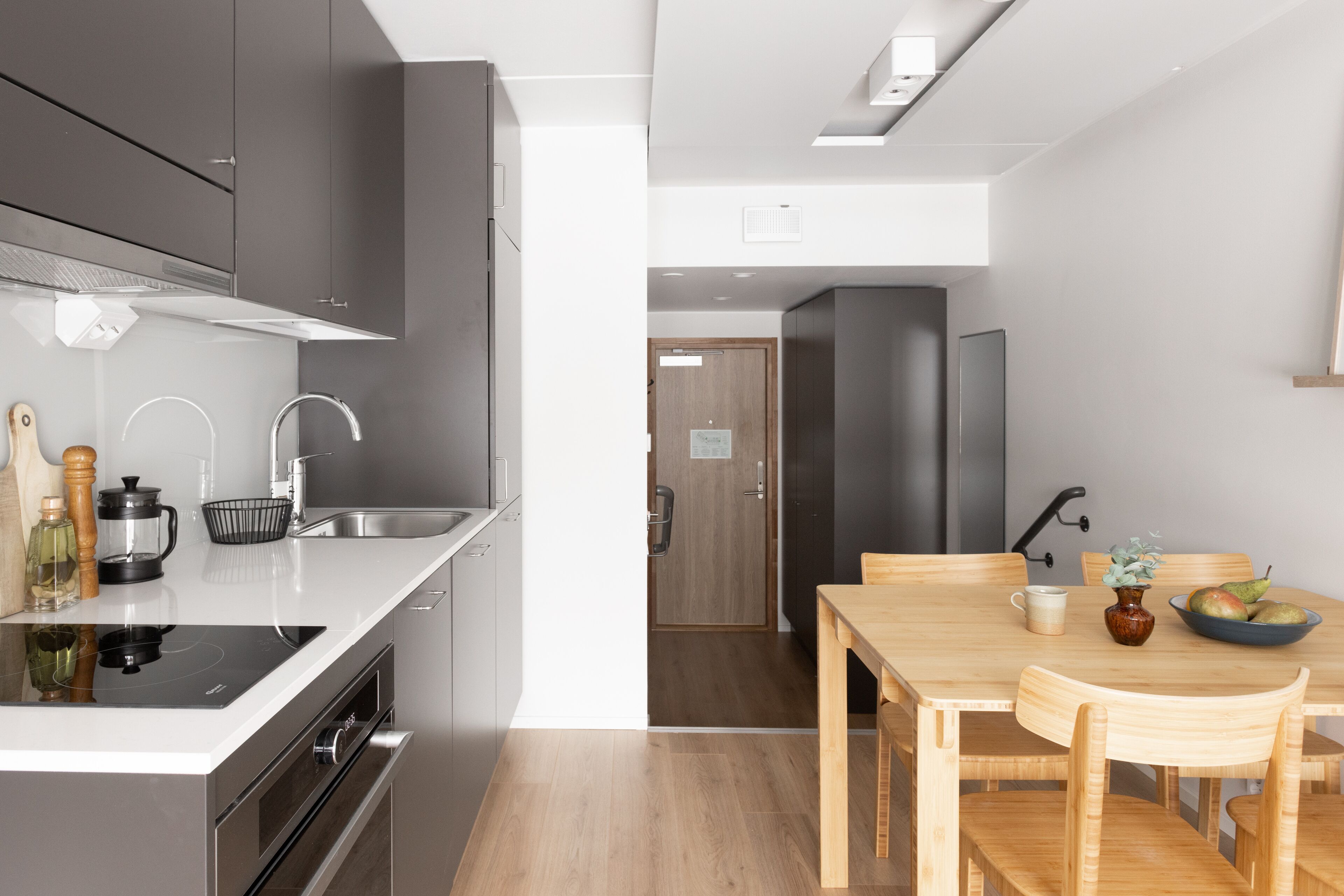 A modern kitchen with a built-in stove, sink, and dark cabinets. There is a dining area with a wooden table and chairs, and a small vase with flowers on the table.