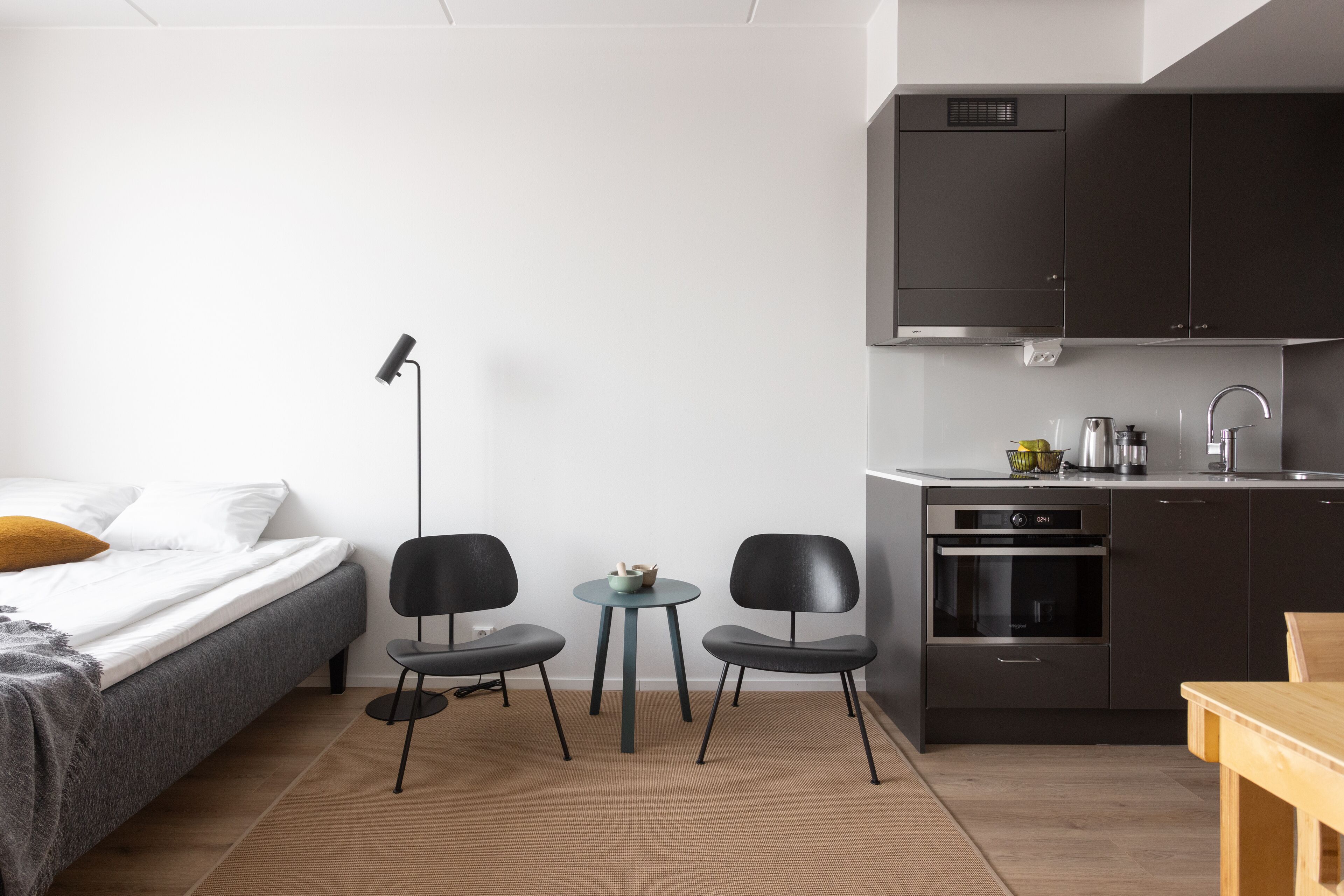 Een kleine kamer met een bed, een eettafeltje, twee stoelen, een kitchenette en een vloerlamp.