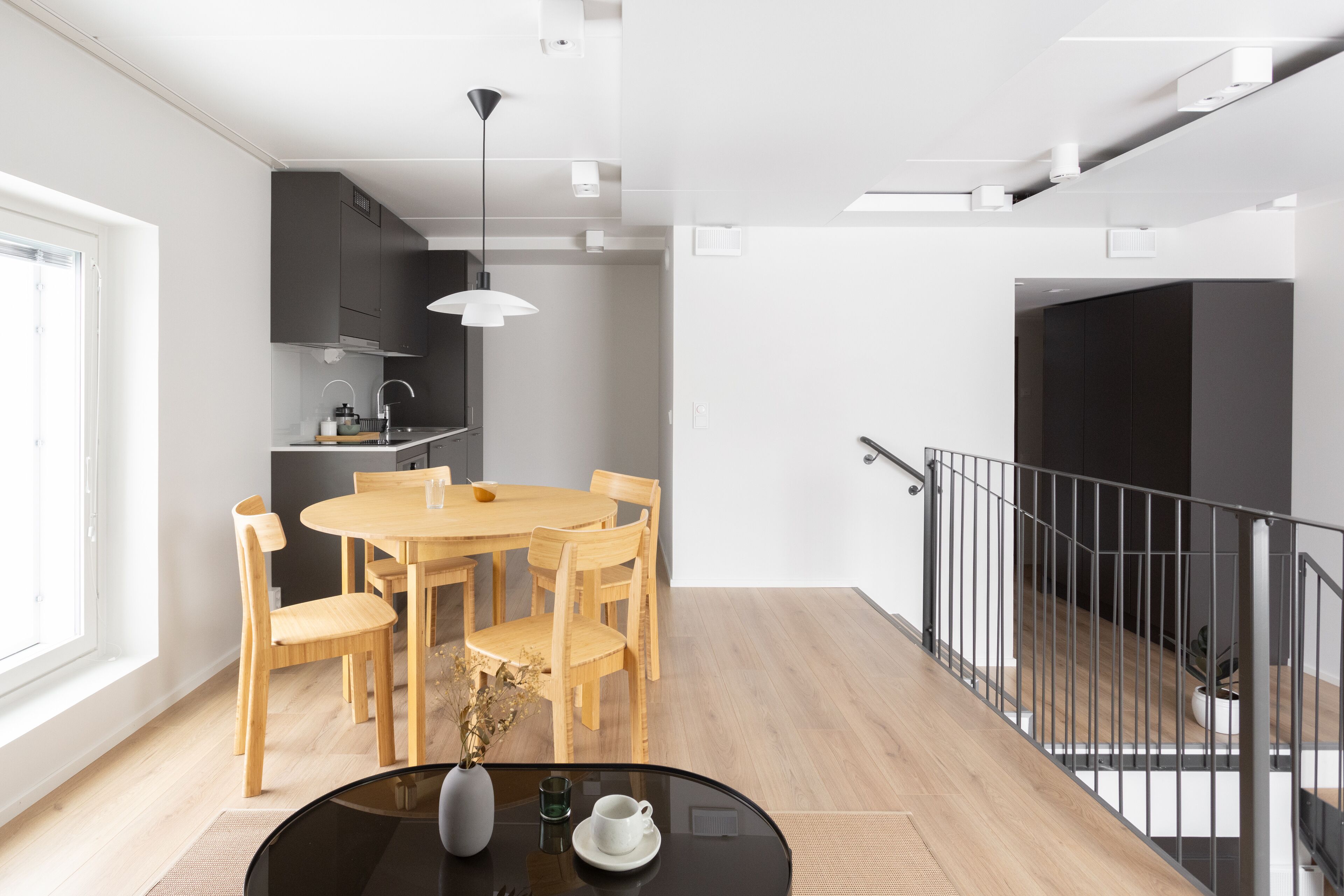 A modern kitchen with dark cabinets, a wooden dining table, and a black railing on the staircase.