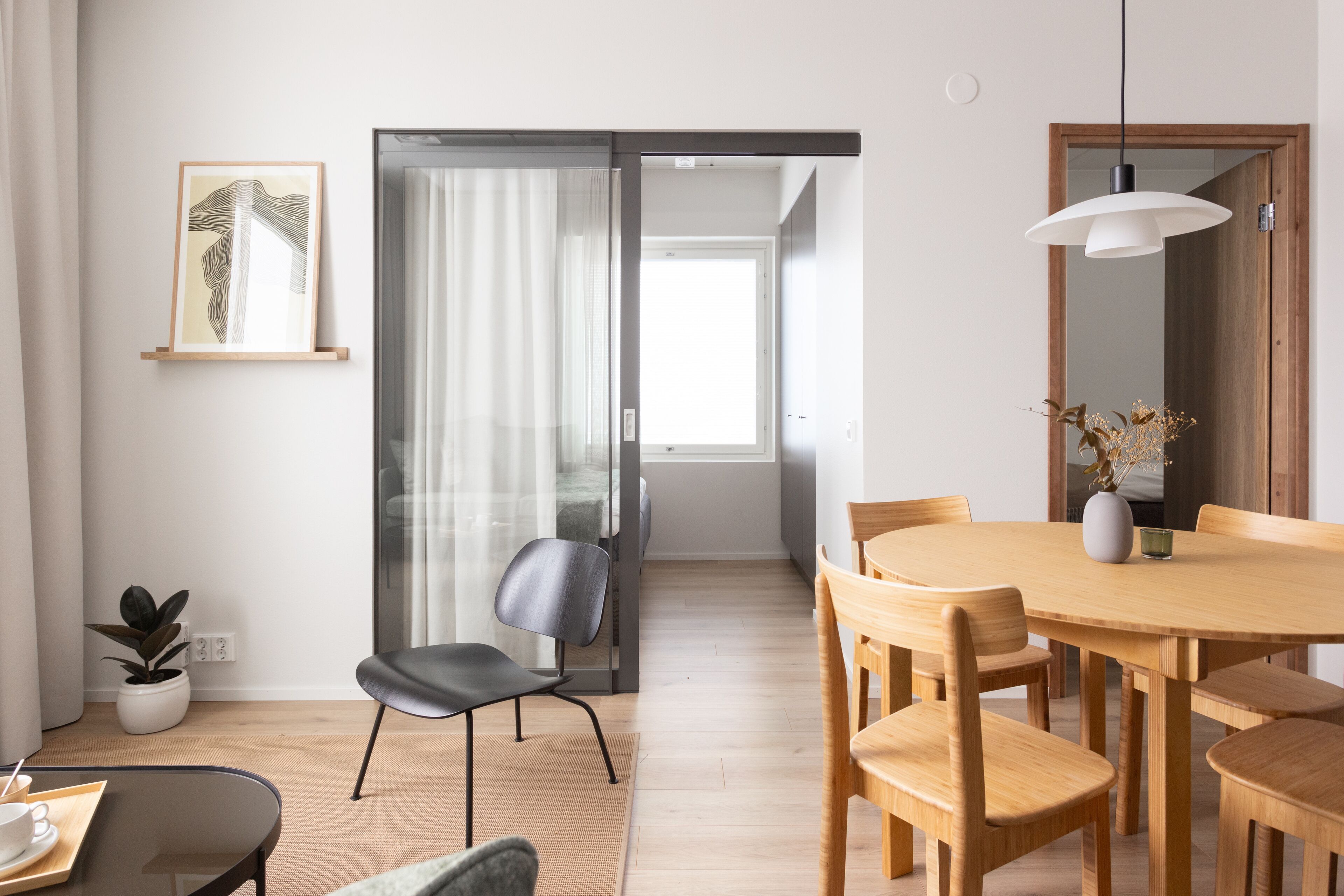 Een moderne woonkamer met een houten eettafel, zwarte stoelen, een glazen scheidingswand, een potplant en een lijstwerk aan de muur.