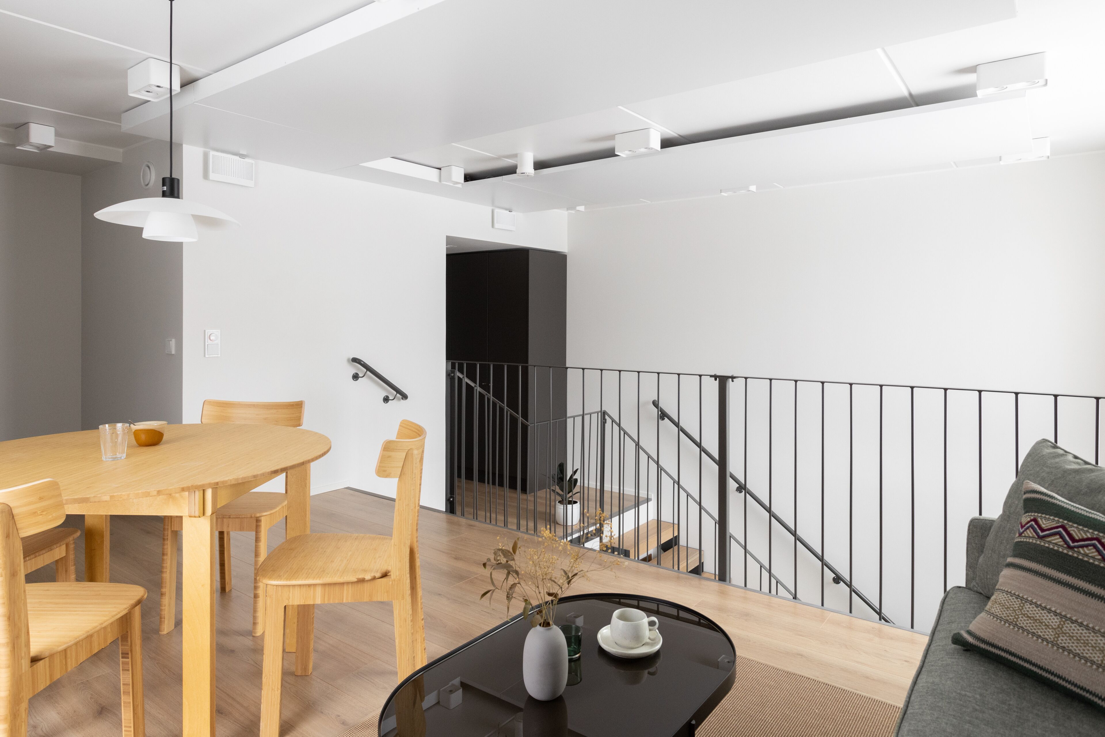 A modern living space with a dining area featuring a wooden table and chairs, a black coffee table with a vase and cup, and a staircase with a metal railing.