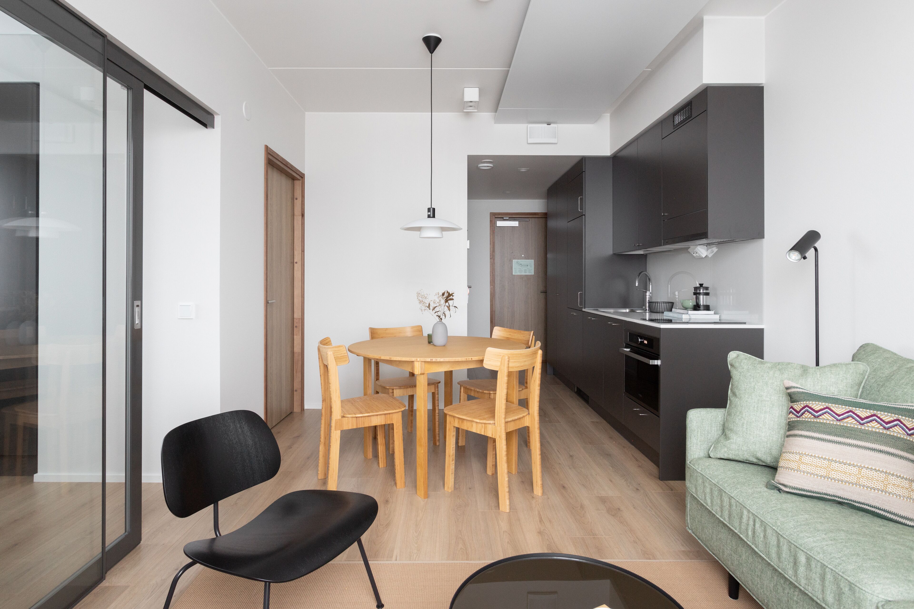 A modern kitchen with dark cabinets, a dining area with a round table and chairs, and a green sofa with a patterned pillow.