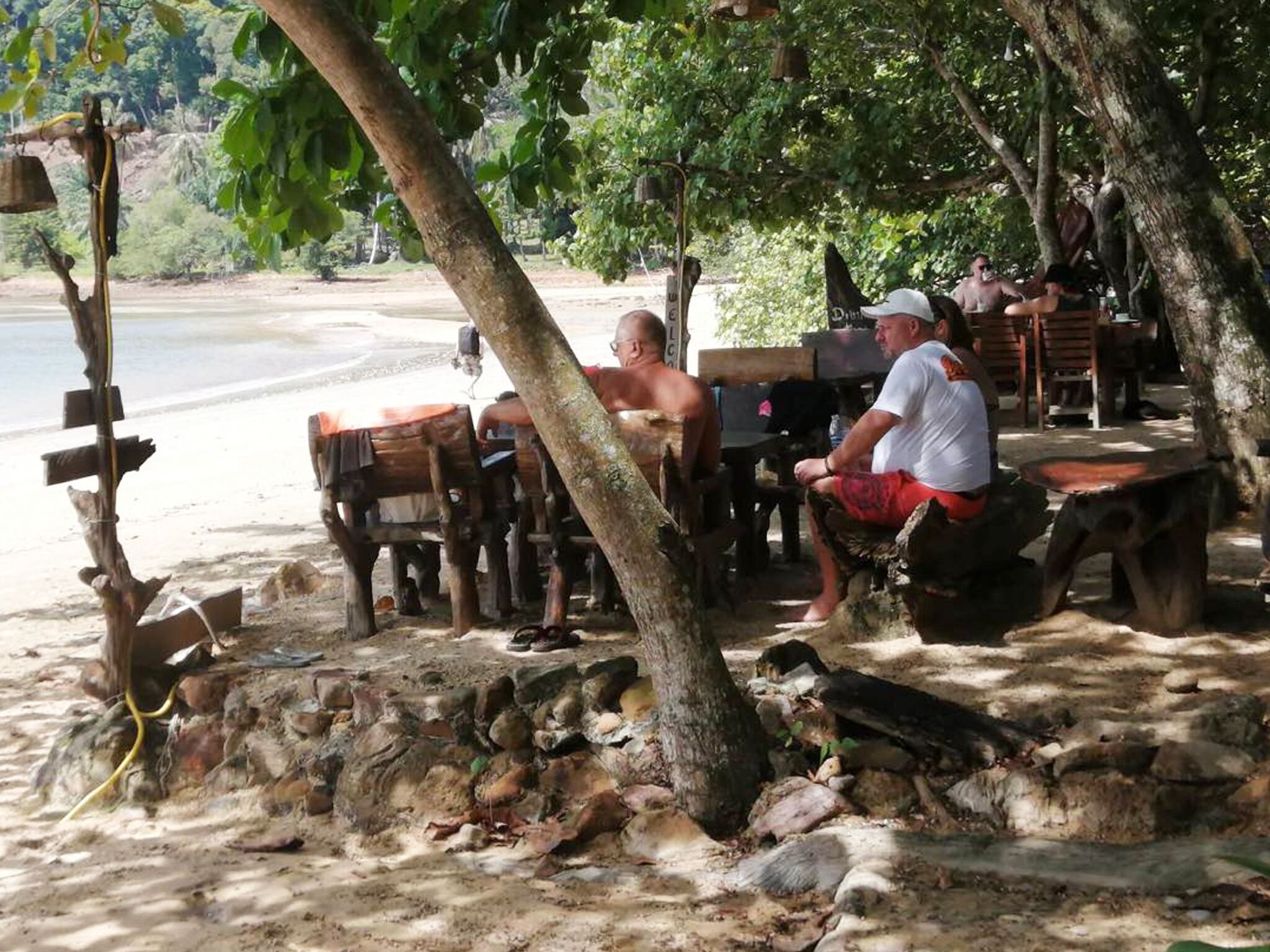 Ein Essbereich am Strand mit Holztischen und -bänken, umgeben von Bäumen und einem felsigen Vordergrund.