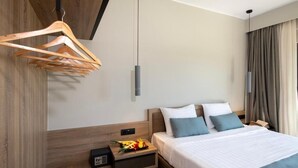 A modern hotel room with a wooden headboard, a bed with white and blue bedding, a bedside table with a fruit basket, and a wooden hanger rack. - Mare Garden (Apokoronas)