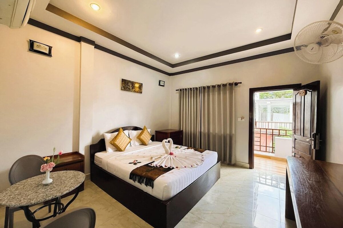 A hotel room with a bed, a small table, a chair, a fan, and a view of the outside.