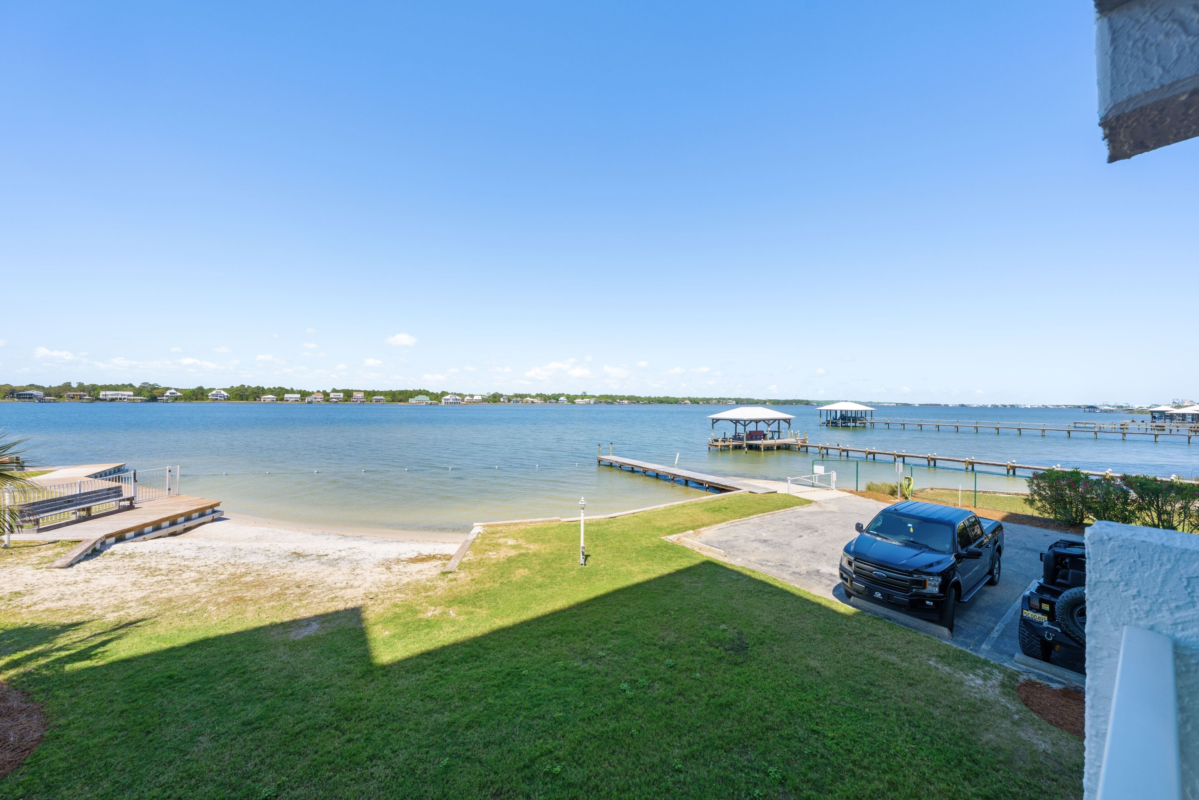A view of a lake with a dock, a boat, and a parking area with a vehicle.