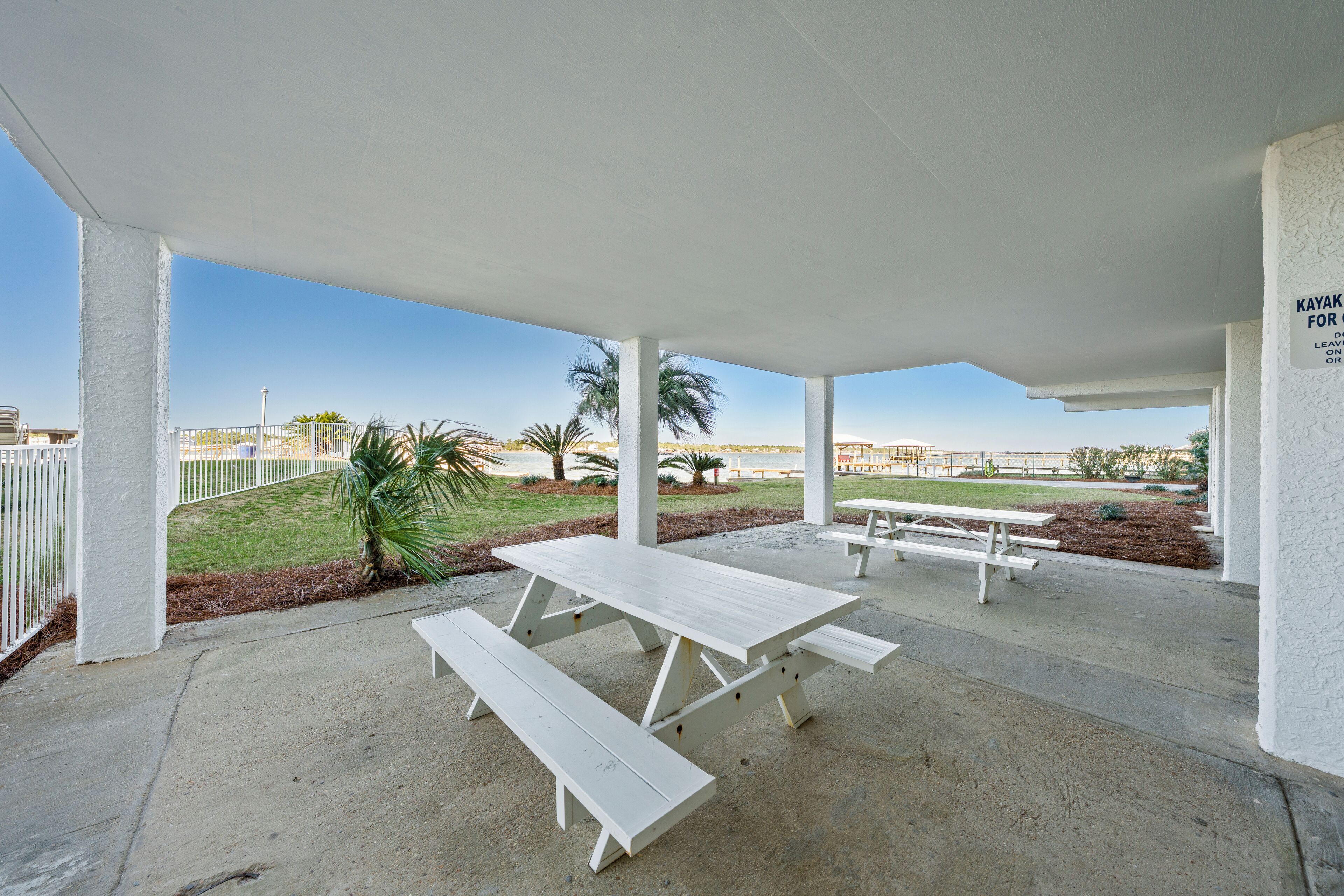 An outdoor covered picnic area with white benches and picnic tables, surrounded by palm trees and a clear sky.