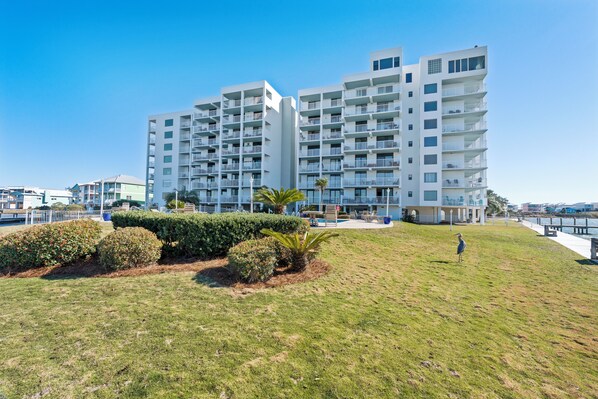 A modern multi-story hotel with balconies, a grassy lawn, and a clear view of the waterfront. - Compass Point 108 (Gulf Shores)