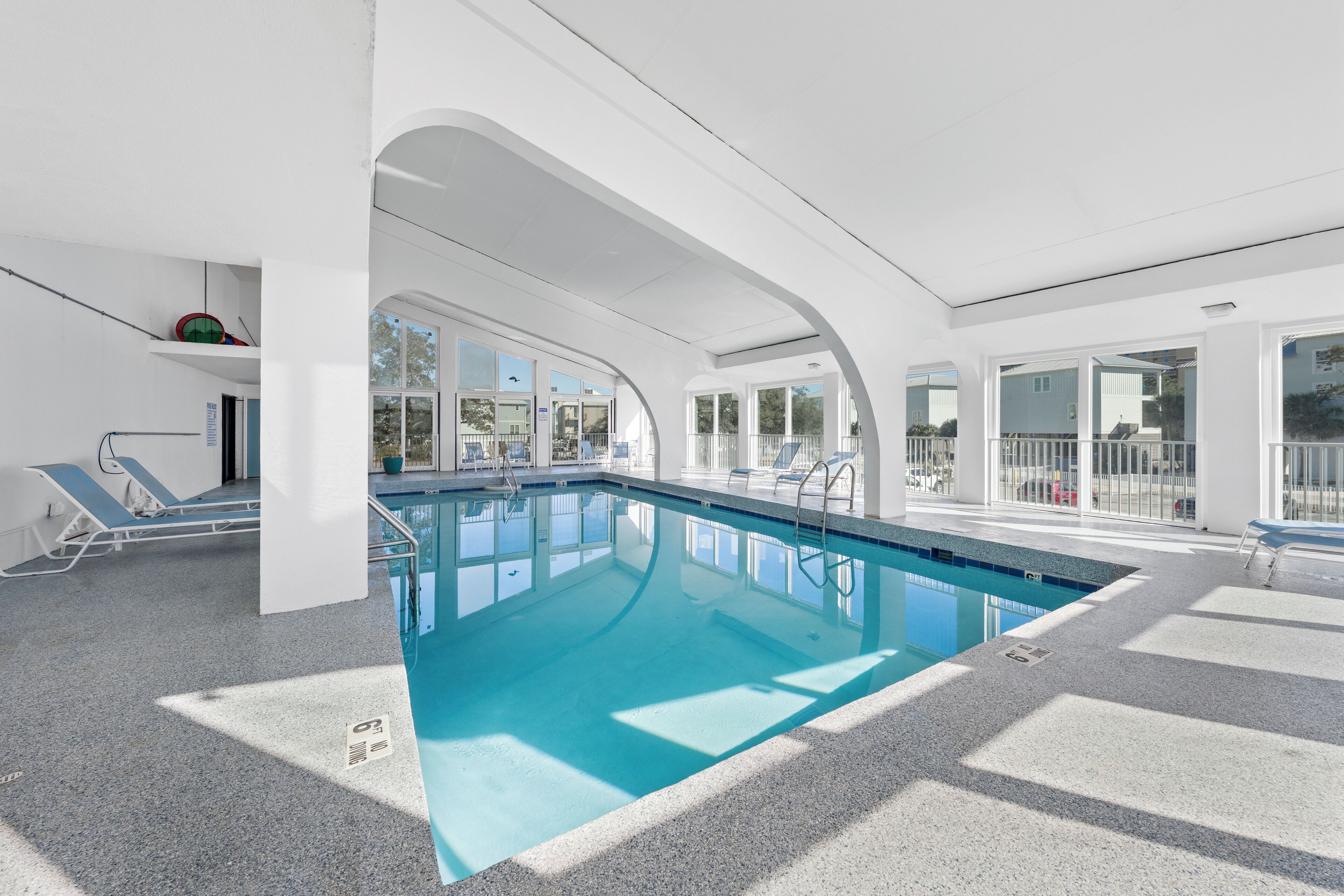 An indoor swimming pool with clear blue water, surrounded by white walls and large windows.