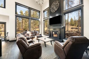 A modern living room with a fireplace, leather sofas, and large windows offering a forest view. - New Misty Pine - Hot tub, Fireplace, AC, Garage (Lead)
