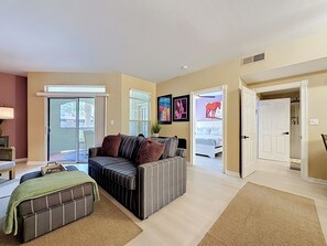 A living room with a sofa, a coffee table, a green throw blanket, a sliding glass door, a bed with a horse decoration, and a hallway leading to a bedroom.