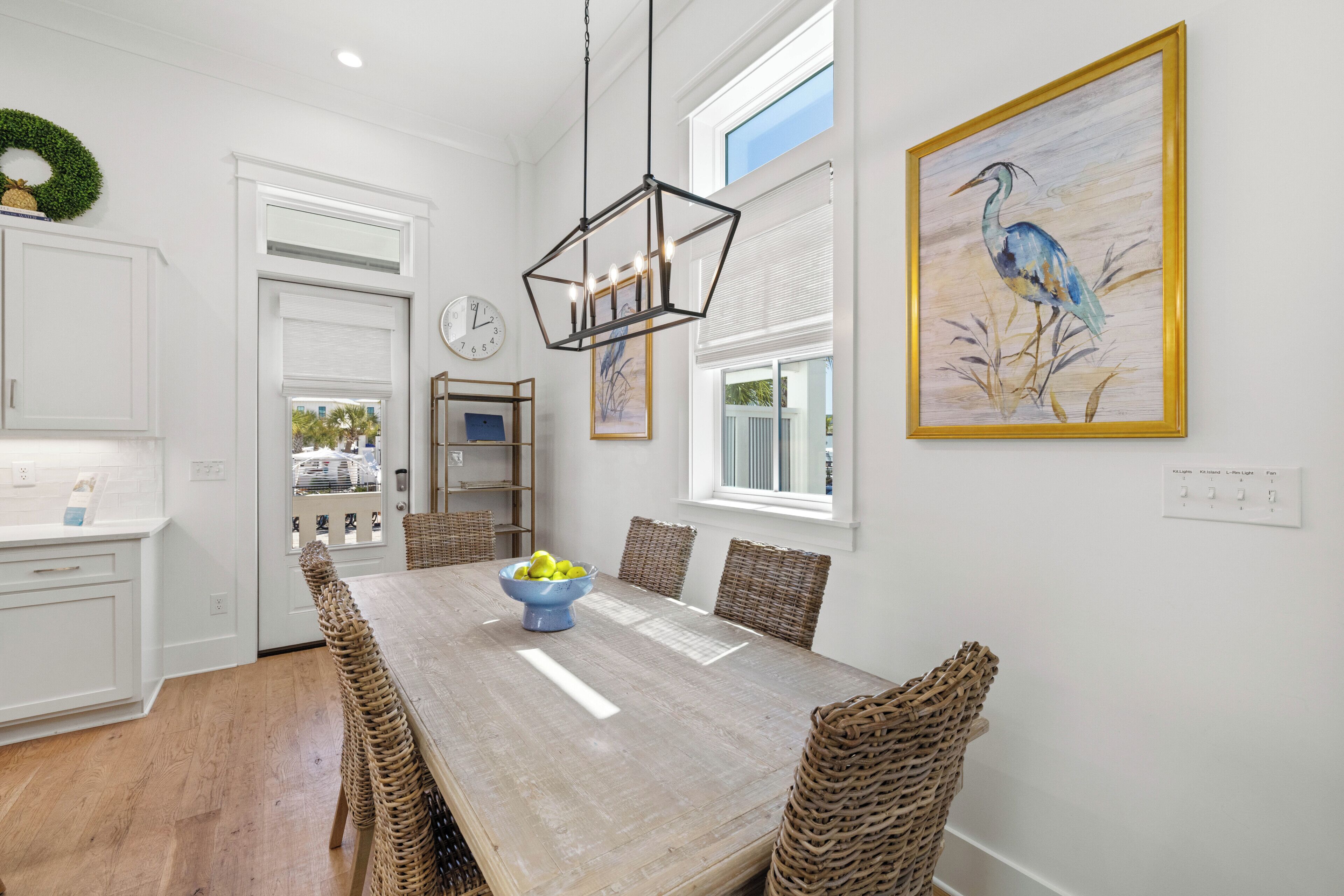 A dining area with a wooden table, wicker chairs, a blue bowl with fruit, and a wall-mounted painting of a bird.