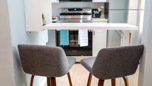 Una cocina moderna con gabinetes blancos, un microondas de acero inoxidable y un horno empotrado. Hay dos sillas tapizadas en gris frente a la encimera.