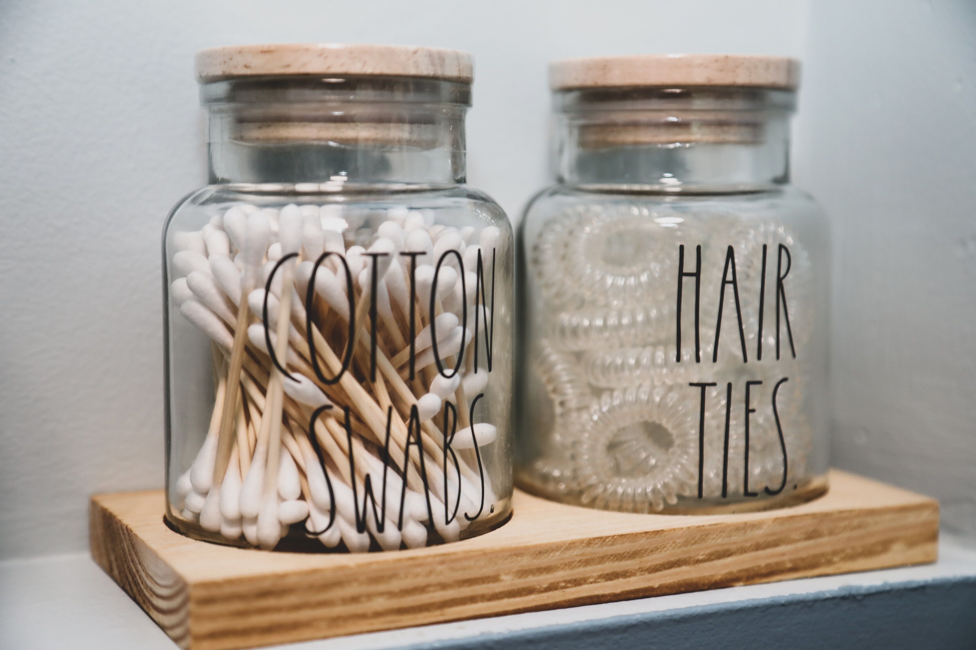 Two glass jars with wooden lids, one labeled "Cotton Swabs" and the other "Hair Ties," placed on a wooden shelf.