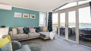 A modern living room with a grey sectional sofa, a small white table, and a grey rug. There are three framed pictures on the teal wall, and a sliding glass door leading to an outdoor area with a view of a body of water. - Lake View Lodge (Bedford)