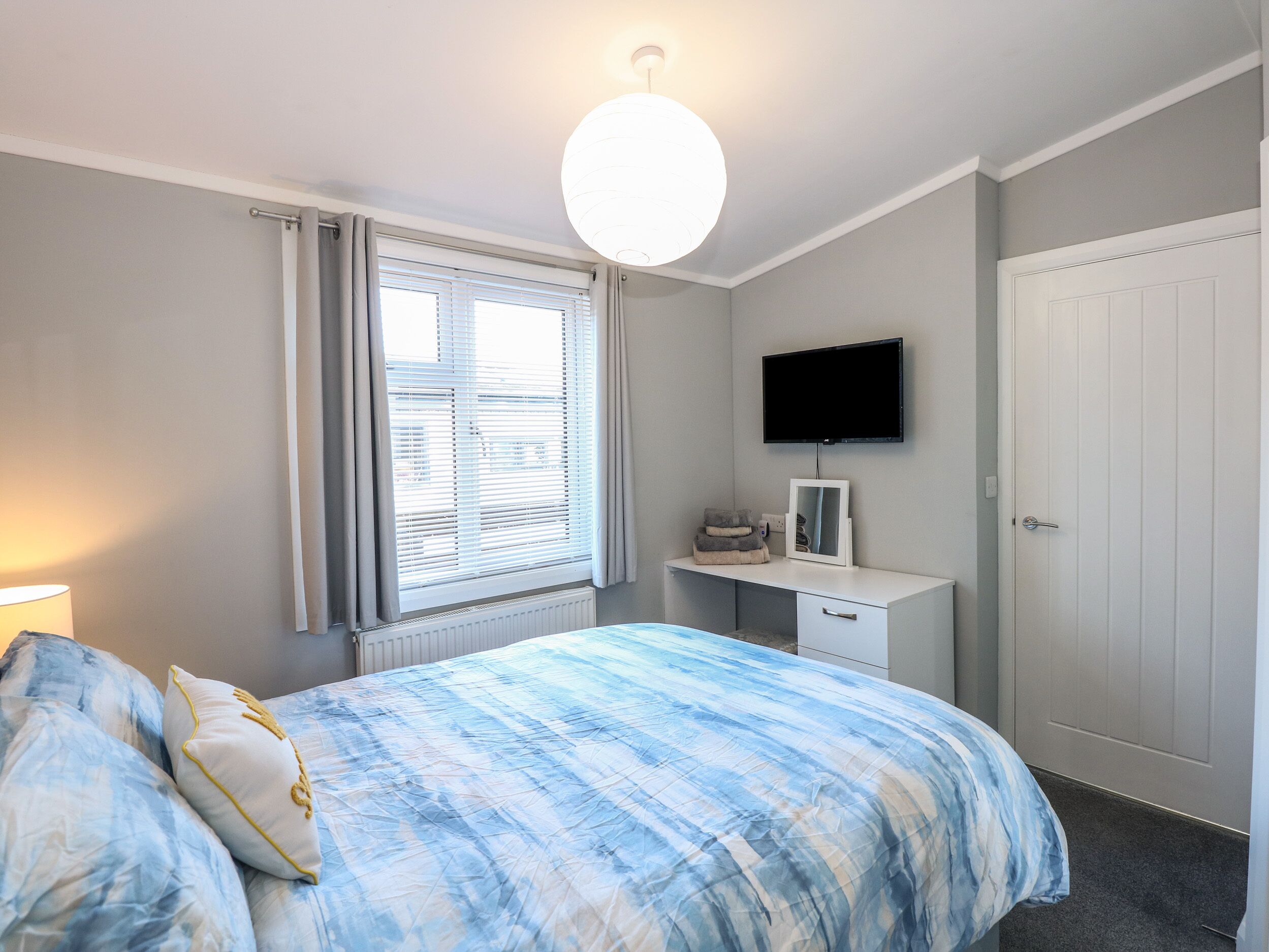 A bedroom with a bed, a bedside table, a TV mounted on the wall, and a window with blinds.