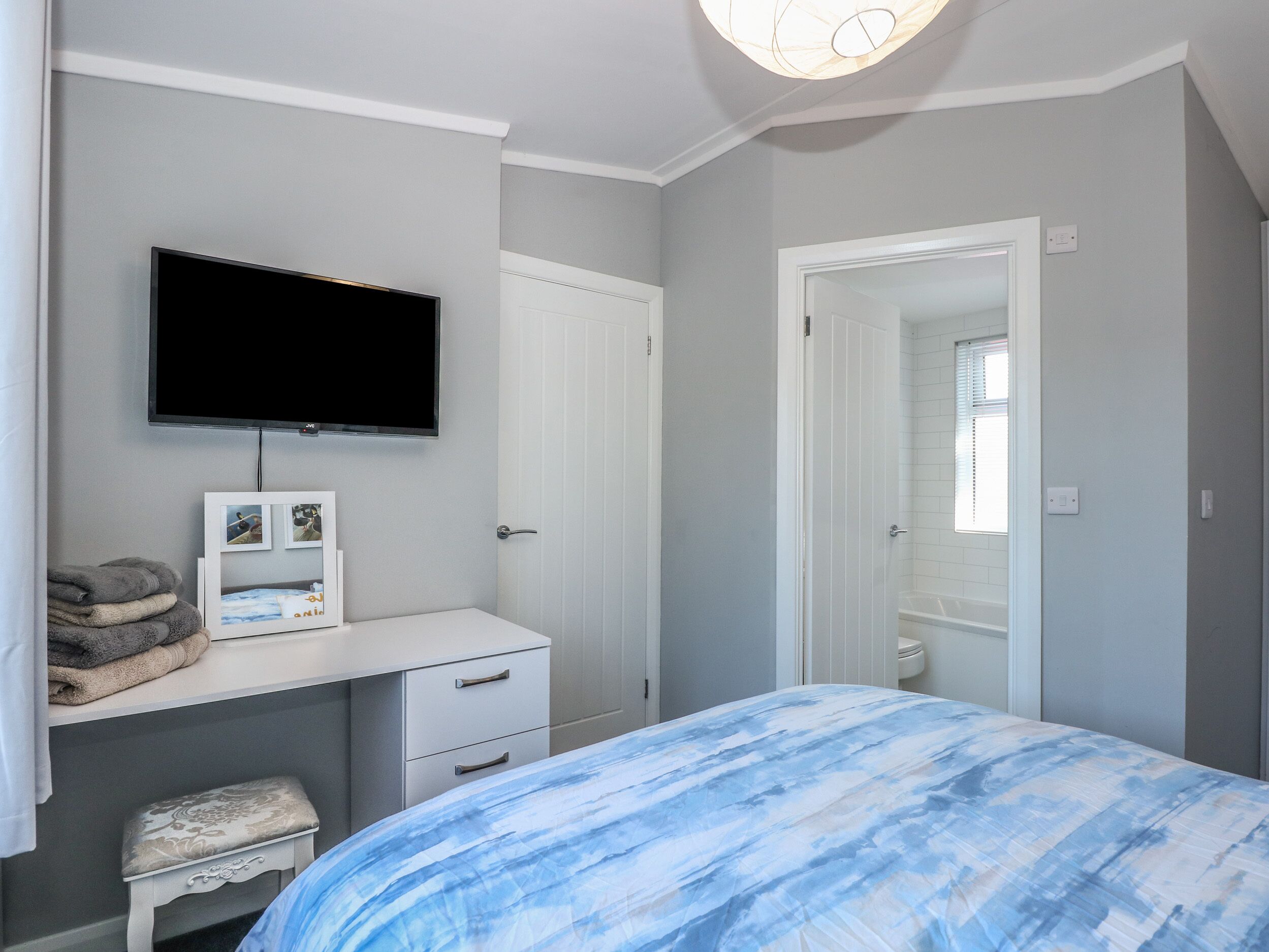 A bedroom with a bed, a TV mounted on the wall, a desk with drawers, and a door leading to a bathroom.