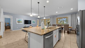Een moderne keuken met een centraal eiland, roestvrijstalen apparatuur, hangende verlichting en een eetgedeelte met uitzicht op de omgeving.