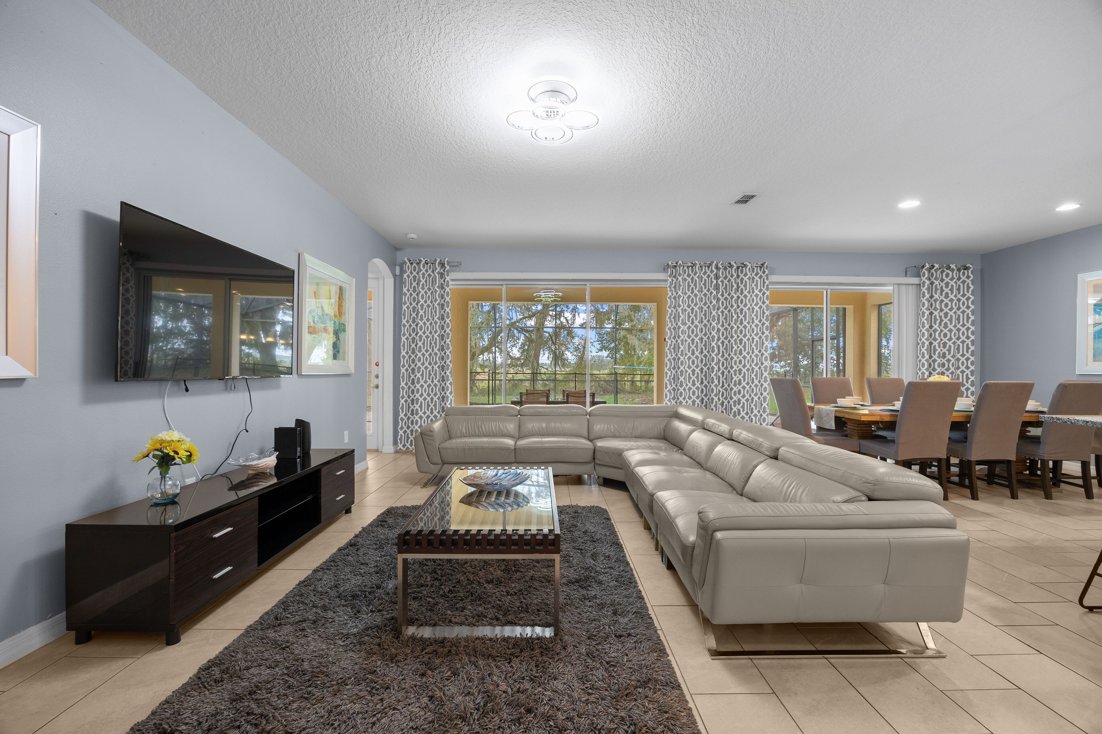 A modern living room with a sectional sofa, a glass coffee table, a dark wooden TV stand, and a large window with patterned curtains.