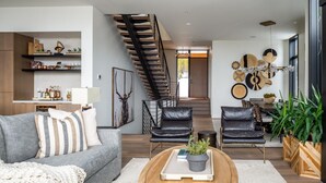 Een moderne woonkamer met een grijze bank, zwarte lederen fauteuils, een houten salontafel en een trap naar een hoger niveau.