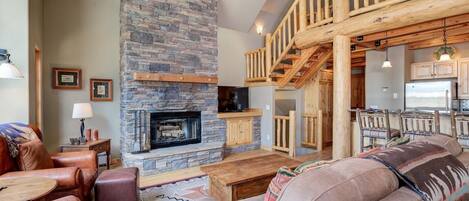 A cozy living room with a stone fireplace, leather furniture, and a wooden staircase leading to a kitchen.