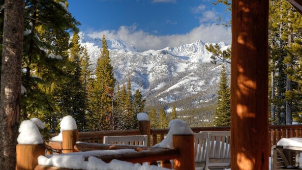 Une terrasse en bois aux garde-corps recouverts de neige, offrant une vue sur un paysage montagneux où les arbres sont également couverts de neige et le ciel est dégagé.
