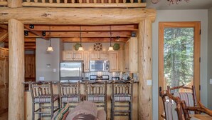 Une cuisine rustique avec des poutres en bois, un îlot central doté de tabourets de bar et une grande fenêtre donnant sur la forêt.