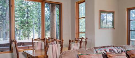 A cozy room with a dining table and chairs, large windows with a forest view, and a sofa with patterned cushions.