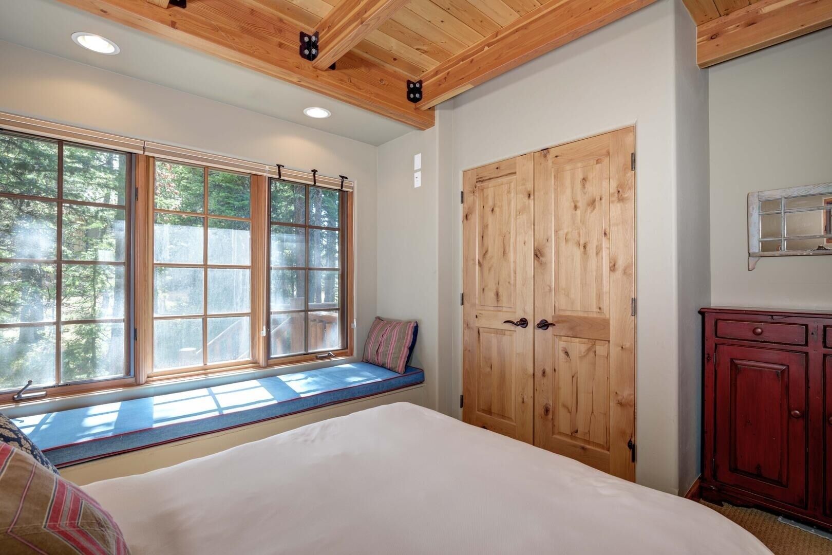Ein Schlafzimmer mit einem großen Bett, einer Sitzbank am Fenster, einem Holzschrank und Blick auf Bäume.