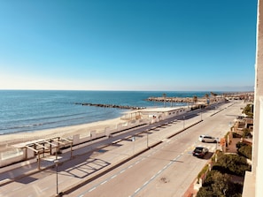 A coastal road with a beach on one side, a clear sky, and a parking area with a few cars.