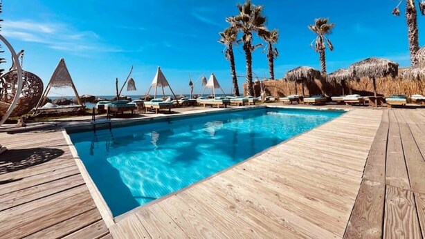 Une terrasse en bois avec une piscine, des chaises longues, des palmiers et des parasols en feuilles de palmier.