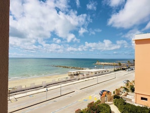 Une route côtière bordée d’une plage et avec l’océan en toile de fond, un espace de stationnement avec des places délimitées, et un bâtiment doté d’un balcon offrant une vue panoramique.