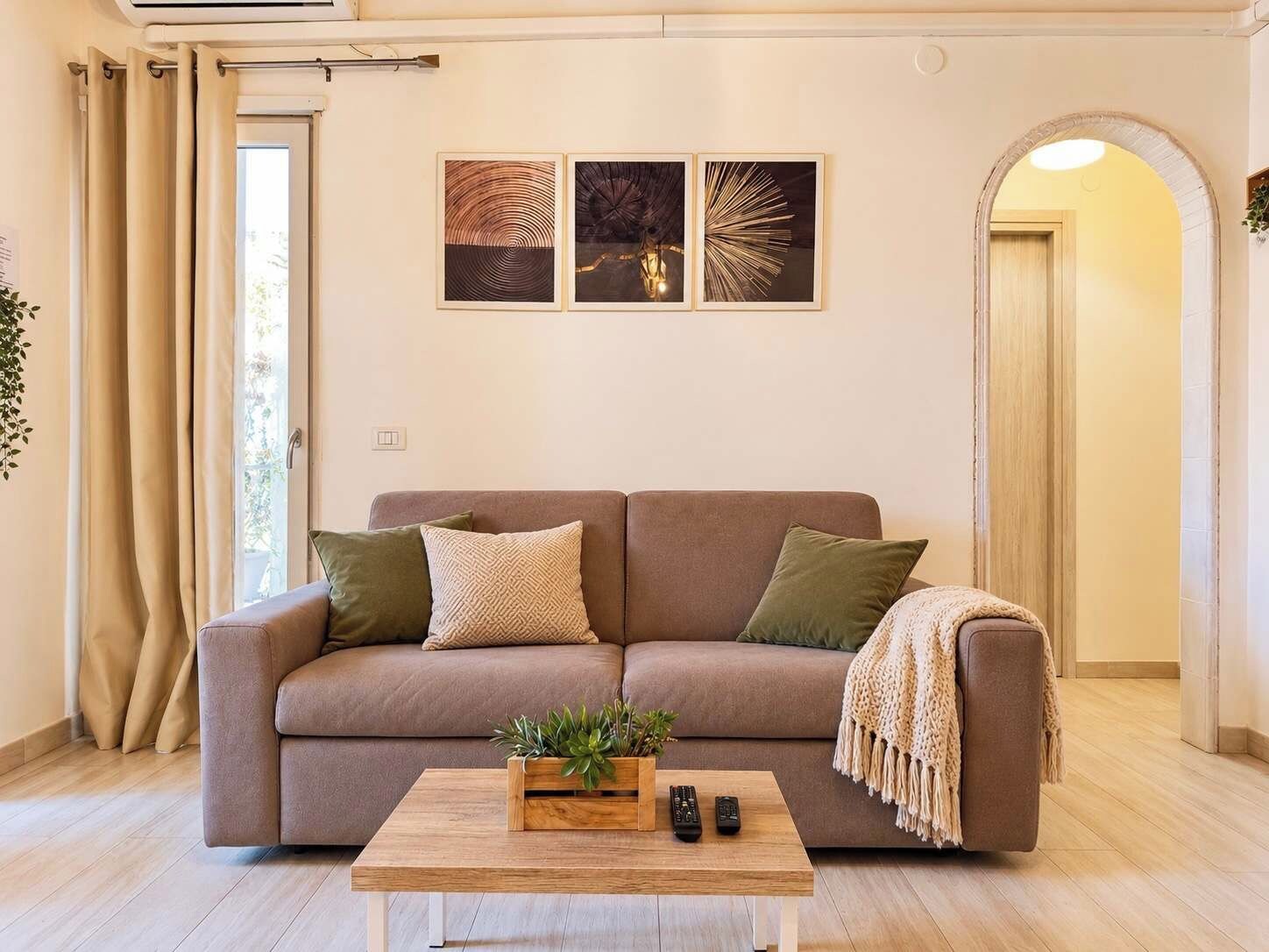 A living room with a sofa, a coffee table with a plant and remotes, and three framed artworks on the wall.