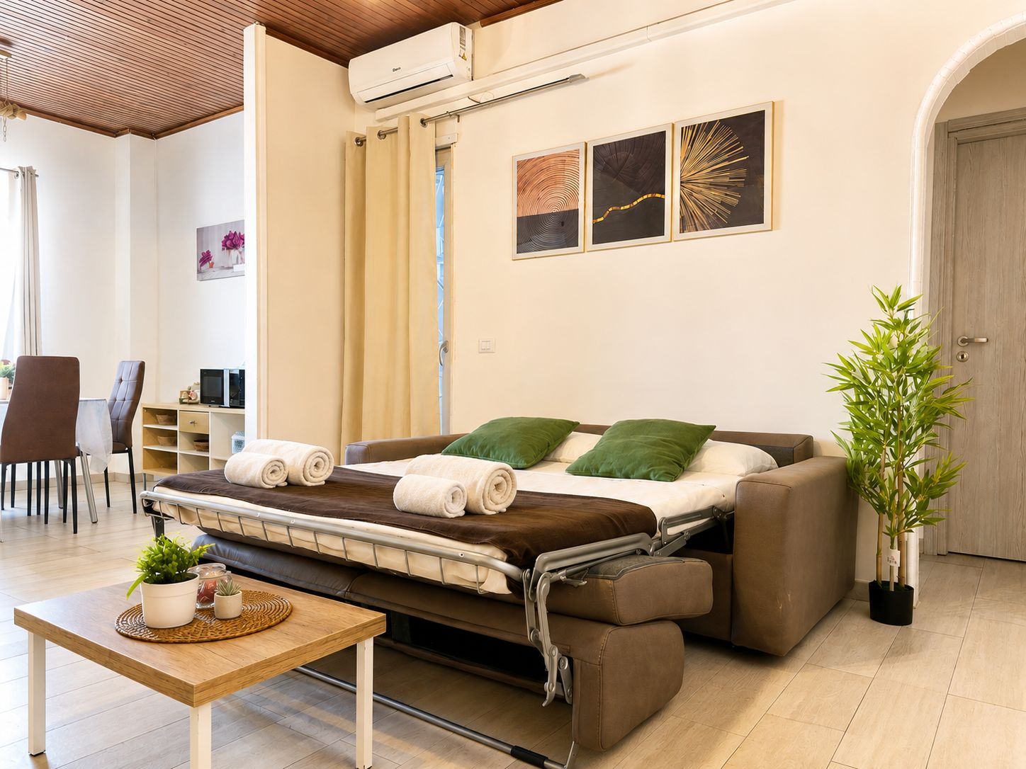 A convertible sofa bed with a table and chairs in a room with a wooden ceiling, a wall-mounted air conditioner, and a potted plant.