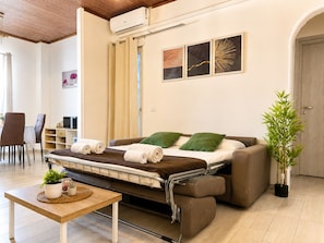 A convertible sofa bed with a table and chairs in a room with a wooden ceiling, a wall-mounted air conditioner, and a potted plant.