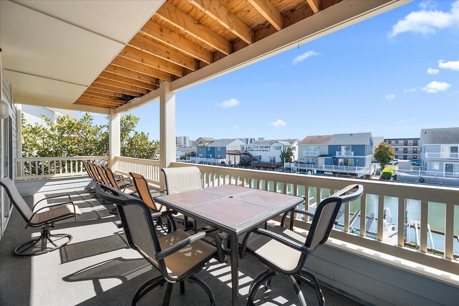 A covered outdoor seating area with a table and chairs, overlooking a waterfront with buildings in the background.