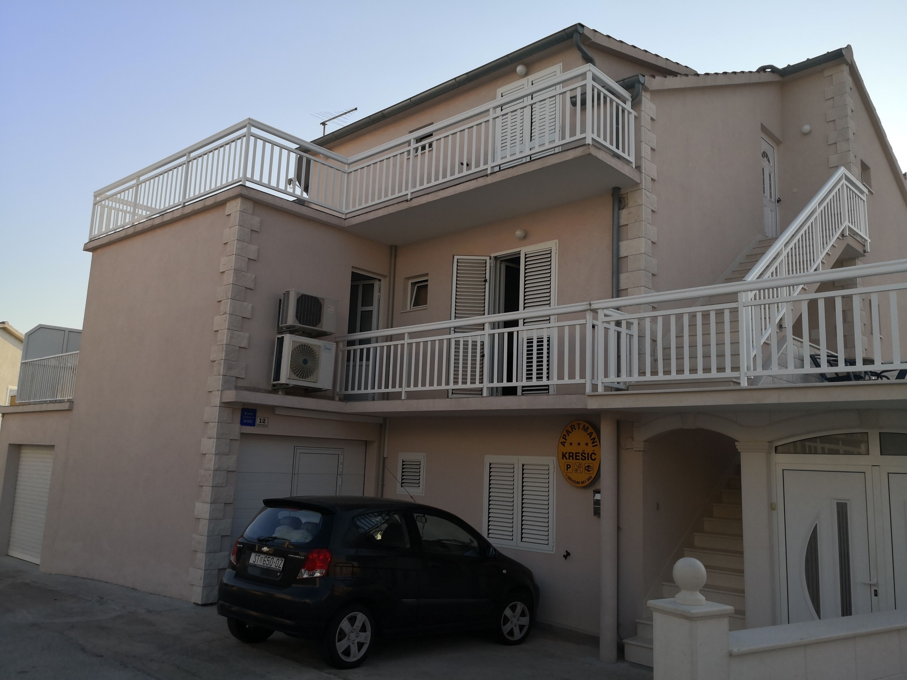 A multi-story building with balconies, a car parked in front, and a sign indicating a parking area.