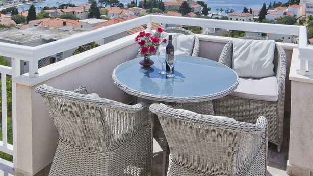 A balcony with wicker furniture, a table with a vase of flowers, and a view of the sea and distant buildings. - Apartmani Kresic 1 (Hvar)